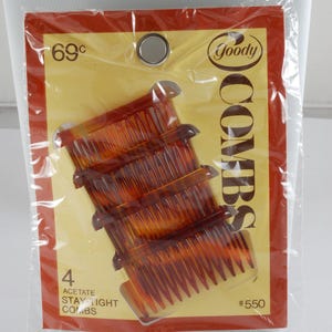 May include: A package of four acetate combs, brand name Goody, in a clear plastic wrapper. The combs are a tortoiseshell color. The package has a red border and a yellow background with the text "4 Acetate Stay Tight Combs".