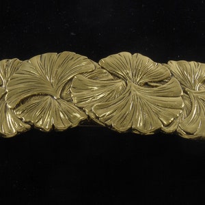 May include: A gold-colored metal barrette featuring a repeating pattern of stylized ginkgo leaves. The leaves are intricately detailed with visible veins and a textured surface, set against a dark background.
