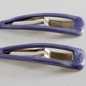 Könnte beinhalten: Zwei lilafarbene Haarklammern aus Kunststoff mit silbernen Metallverschlüssen. Die Klammern haben ein marmoriertes Muster.
