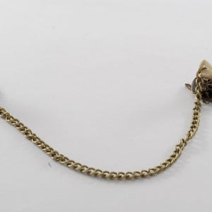 May include: A pair of gold-toned metal garment clips connected by a chain. The clips have a decorative design.