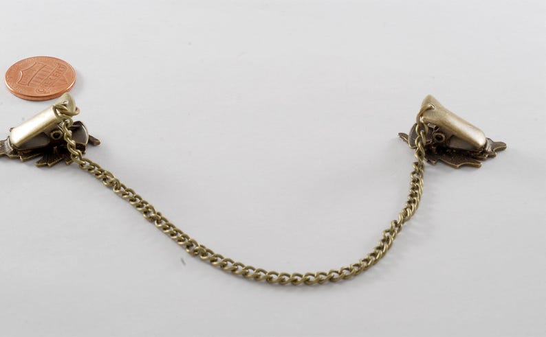 May include: A pair of gold-toned metal garment clips connected by a chain. The clips have a decorative design.