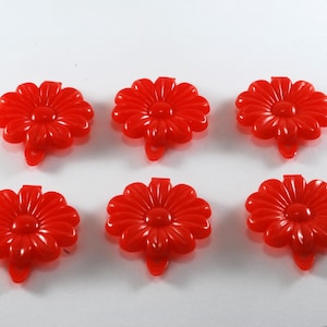 May include: Six red plastic flower-shaped buttons with a raised center. The buttons have a small loop on the back for attaching to fabric.
