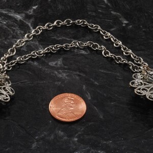May include: A pair of silver filigree brooches connected by a silver chain. The brooches are in the shape of a flower with a pin on the back. The chain is about the size of a US penny.