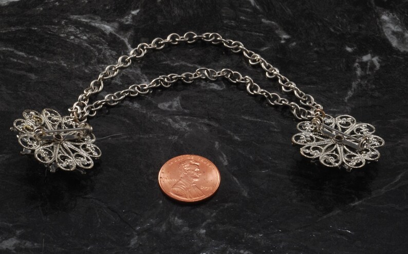 May include: A pair of silver filigree brooches connected by a silver chain. The brooches are in the shape of a flower with a pin on the back. The chain is about the size of a US penny.
