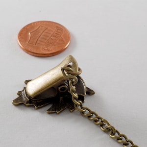 May include: A bronze-coloured metal clasp with a decorative floral design and a chain attached. The clasp is about the size of a UK penny.