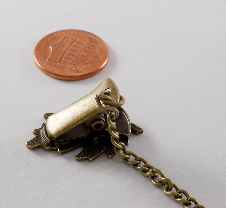 May include: A bronze-coloured metal clasp with a decorative floral design and a chain attached. The clasp is about the size of a UK penny.