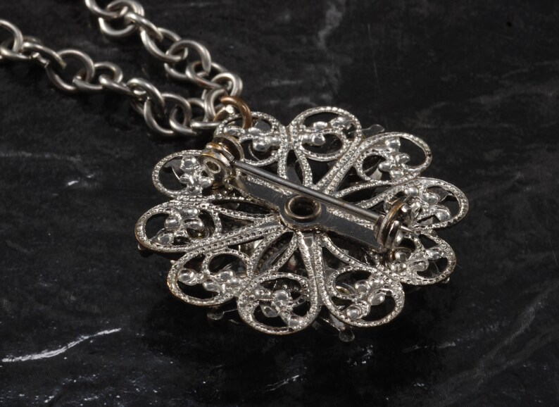 May include: Silver filigree pendant with a pin back closure. The pendant is in the shape of a flower with a delicate design.