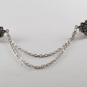 Puede incluir: Un broche de doble cadena de tono plateado con dise&ntilde;os florales ornamentados y diamantes de imitaci&oacute;n transparentes.