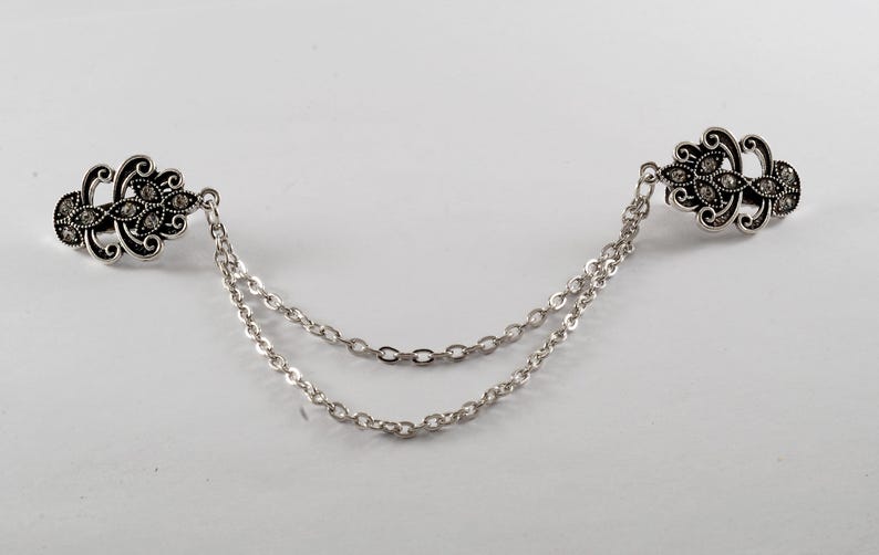 Puede incluir: Un broche de doble cadena de tono plateado con dise&ntilde;os florales ornamentados y diamantes de imitaci&oacute;n transparentes.