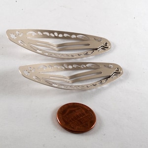 K&ouml;nnte beinhalten: Zwei silberfarbene Haarspangen mit dekorativen Aussparungen. Eine M&uuml;nze liegt neben den Spangen zum Gr&ouml;&szlig;envergleich. Die Spangen sind aus Metall und haben einen Federmechanismus.