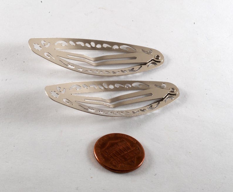 K&ouml;nnte beinhalten: Zwei silberfarbene Haarspangen mit dekorativen Aussparungen. Eine M&uuml;nze liegt neben den Spangen zum Gr&ouml;&szlig;envergleich. Die Spangen sind aus Metall und haben einen Federmechanismus.