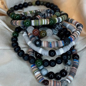 Op de afbeelding: Een stapel armbanden met zwarte, groene, grijze en witte kralen. De armbanden zijn gemaakt van verschillende materialen, waaronder hout, steen en metaal.