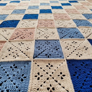 Puede incluir: Una manta de ganchillo con un patrón de cuadros en tonos de azul, beige y rosa. La manta está hecha de un hilo suave y texturizado y tiene un diseño delicado y de encaje.