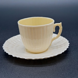 Puede incluir: Juego de taza y platillo de color crema. La taza tiene un diseño acanalado y un asa curvada. El platillo tiene un borde festoneado. El interior de la taza es de color amarillo pálido. El juego es de cerámica.