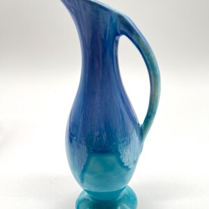 May include: A small blue and turquoise ceramic pitcher with a curved handle. The pitcher has a glazed finish and a rounded base.