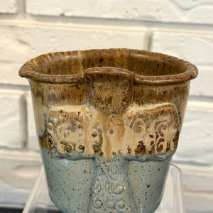 May include: Handmade ceramic pot with a unique design. The top and handles are brown, transitioning to a light tan and blue base. The pot features embossed elephant-like figures and decorative swirls. A rustic, artistic piece.