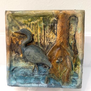 Könnte beinhalten: Quadratisches, strukturiertes Kunstwerk mit einem Relief eines Reihers im Wasser in der Nähe eines Baumes. Das Kunstwerk verwendet Blau-, Grün- und Brauntöne und schafft eine naturalistische Szene. Der Reiher ist grau, und das Wasser ist eine Mischung aus Blau und Grün.