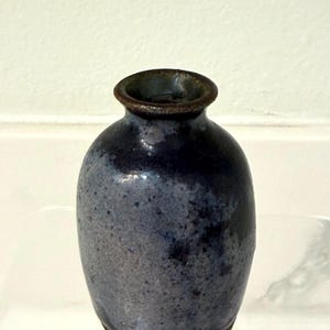 Könnte beinhalten: Kleine, handgefertigte Keramikvase mit dunkelblauer Glasur. Die Vase hat einen abgerundeten Körper und eine schmale Öffnung, mit einer gesprenkelten Textur und einem dunklen Rand. Der Boden ist hellbraun.