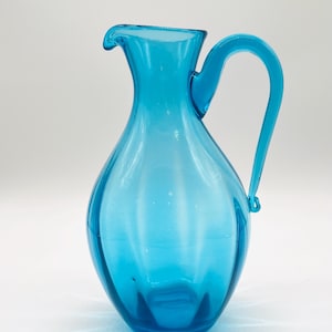 May include: A blue glass pitcher with a rounded body and a curved handle. The pitcher has a textured surface and a wide mouth.