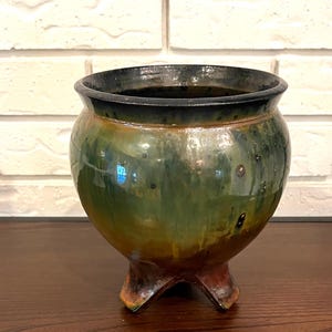 May include: A handcrafted ceramic pot with a glossy, mottled green and brown glaze. The pot has a rounded body, a dark rim, and three small, sturdy feet. The interior is dark, and the pot sits on a wooden surface.