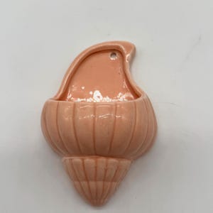 May include: A peach-colored ceramic wall pocket shaped like a seashell. The pocket has a rounded body with vertical ridges and a flared, pointed base. A small opening is at the top for hanging.