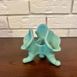 Puede incluir: Un jarrón de cerámica turquesa con un diseño abstracto único. El jarrón tiene una base de tres patas y tres aberturas ensanchadas, con un degradado de tonos azules y verdes. La pieza se exhibe sobre una superficie de madera oscura.
