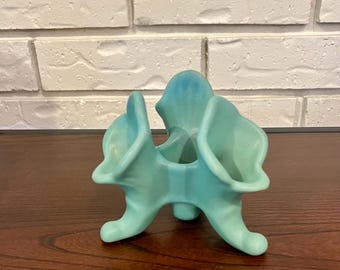 Könnte beinhalten: Eine türkisfarbene Keramikvase mit einem einzigartigen, abstrakten Design. Die Vase hat einen dreibeinigen Sockel und drei ausgestellte Öffnungen mit einem Farbverlauf aus Blau- und Grüntönen. Das Stück wird auf einer dunklen Holzoberfläche präsentiert.