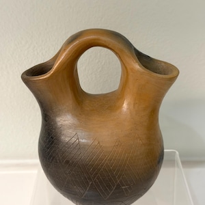 May include: A handcrafted, brown pottery vase with a unique handle and a central opening. The vase features a gradient color scheme, transitioning from light brown to dark brown. Geometric patterns are etched into the lower portion of the vase.