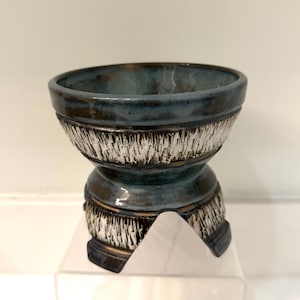 May include: A ceramic bowl with a textured, white and brown band around the middle and base. The top and base are a dark teal colour. The bowl is sitting on a clear acrylic stand.