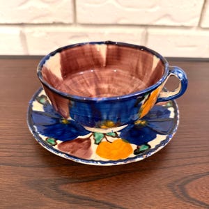 May include: A hand-painted ceramic teacup and saucer set. The cup has a blue rim and a brown interior. The saucer is decorated with blue floral designs, orange accents, and a white border. The set is placed on a wooden surface.