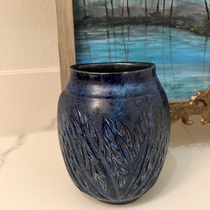May include: A small, dark blue ceramic vase with a textured design of leaves. The vase has a rounded shape and a slightly flared lip.