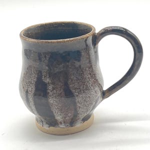 Könnte beinhalten: Eine kleine, braune und weiße Keramiktasse mit abgerundetem Körper und Henkel. Die Tasse hat eine gesprenkelte Glasur und einen leicht ausgestellten Boden.