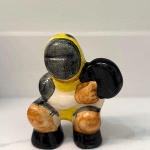 May include: A ceramic figurine of a diver in a yellow and black diving suit. The figurine has a black helmet, black gloves, and brown boots. The diver is in a squatting position.