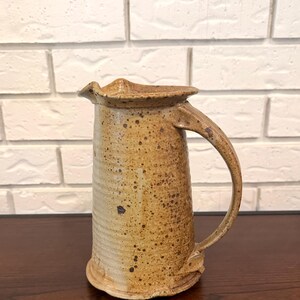 Puede incluir: Jarra de cerámica hecha a mano con un esmalte moteado marrón y beige. La jarra tiene un asa curva y un borde ligeramente ensanchado. La cerámica es de estilo rústico, ideal para servir bebidas.