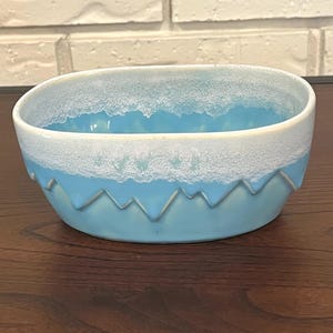 Puede incluir: Cuenco de cerámica ovalado en azul claro con borde blanco. El cuenco tiene un patrón texturizado y ondulado alrededor de la base y el borde. Probablemente para servir o exhibir. Mide unos 20 cm de largo.