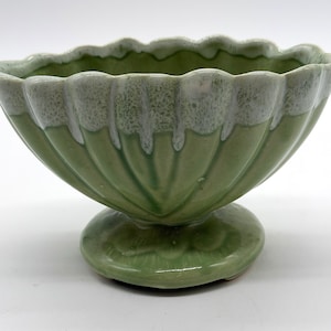 May include: A green ceramic pedestal bowl with a scalloped rim. The bowl has a fluted design and a light green and white glazed finish. The bowl is on a round base. The bowl is likely vintage.