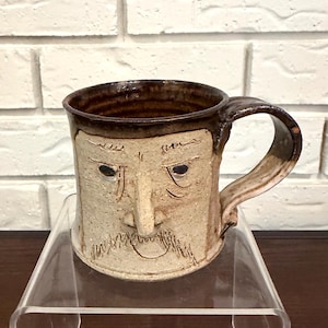 May include: A handmade ceramic mug with a face design. The mug features a light brown base with a darker brown rim and handle. The face has carved details for eyes, a nose, and a beard.