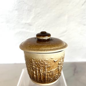 Puede incluir: Un pequeño tarro de cerámica con tapa. El tarro está decorado con un diseño marrón y blanco de árboles y un pájaro. La tapa es marrón con un borde blanco.