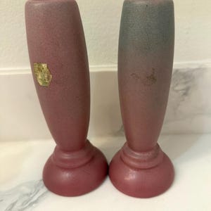 May include: Two vintage bud vases. The vases are a dusty rose color with a darker rose base. One vase has a gold sticker. The tops of the vases are a lighter color, with a gradient effect. The vases are approximately 7 inches tall.