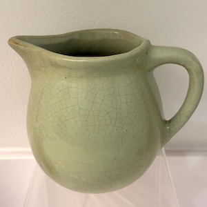 May include: A pale green ceramic pitcher with a rounded body, a curved handle, and a spout. The pitcher has a crackle glaze finish, giving it a textured appearance. The pitcher is displayed on a clear acrylic shelf.