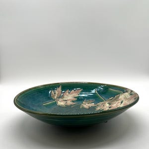 May include: A teal ceramic bowl with a leaf design. The bowl has a wide, shallow shape with a dark green rim. The interior features embossed leaves in shades of pink and gold.