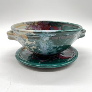May include: A handmade ceramic bowl and saucer set. The bowl is a deep green with blue and red accents, featuring two small handles. The saucer mirrors the bowl's color scheme. This unique set is perfect for serving soups or small dishes.