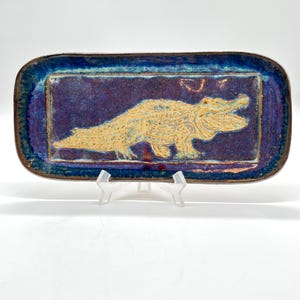 May include: Rectangular ceramic tray with a blue and purple glaze. The center features a raised, textured alligator design in a light tan color. The tray is supported by a clear plastic stand.