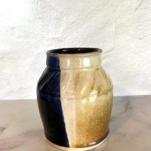 Könnte beinhalten: Eine kleine, handgefertigte Keramikvase mit brauner, weißer und blauer Glasur. Die Vase hat einen runden Körper und eine leicht ausgestellte Lippe.