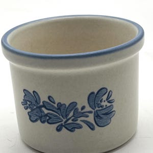 Può includere: Un piccolo contenitore in ceramica bianco sporco con un motivo floreale blu. Il bordo e il motivo floreale sono blu. Il contenitore ha un bordo leggermente angolato e una forma cilindrica. Il design presenta fiori e foglie stilizzati.