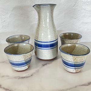 May include: A ceramic sake set featuring a tall, slender pitcher and four small cups. The set has a speckled, off-white glaze with blue horizontal stripes. The cups and pitcher have a rustic, handmade appearance.