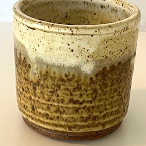 May include: A ceramic planter with a speckled white glaze and a brown glaze with a textured finish. The planter has a rounded bottom and a slightly flared top.