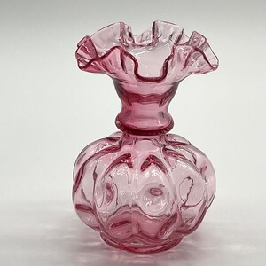Puede incluir: Un jarrón de vidrio rosa con un borde superior con volantes y un cuerpo texturizado y burbujeante. El jarrón mide aproximadamente 15 cm de alto.