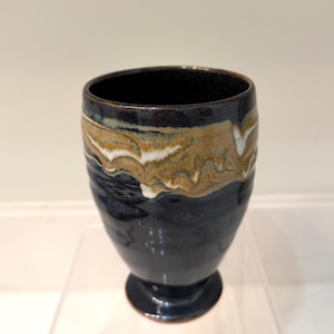 Könnte beinhalten: Eine handgefertigte Keramiktasse mit dunkelblauem Boden und einem dekorativen Band aus beige, weißen und braunen Wirbeln. Die Tasse hat einen Sockel und eine glatte, glänzende Oberfläche. Der Innenraum ist dunkelblau.