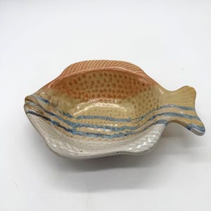 Puede incluir: Un plato de cerámica con forma de pez y superficie texturizada. El plato presenta una paleta de colores beige, naranja y azul, con un borde blanco. El pez tiene un diseño detallado de aleta y cola, perfecto para servir aperitivos o como pieza decorativa.